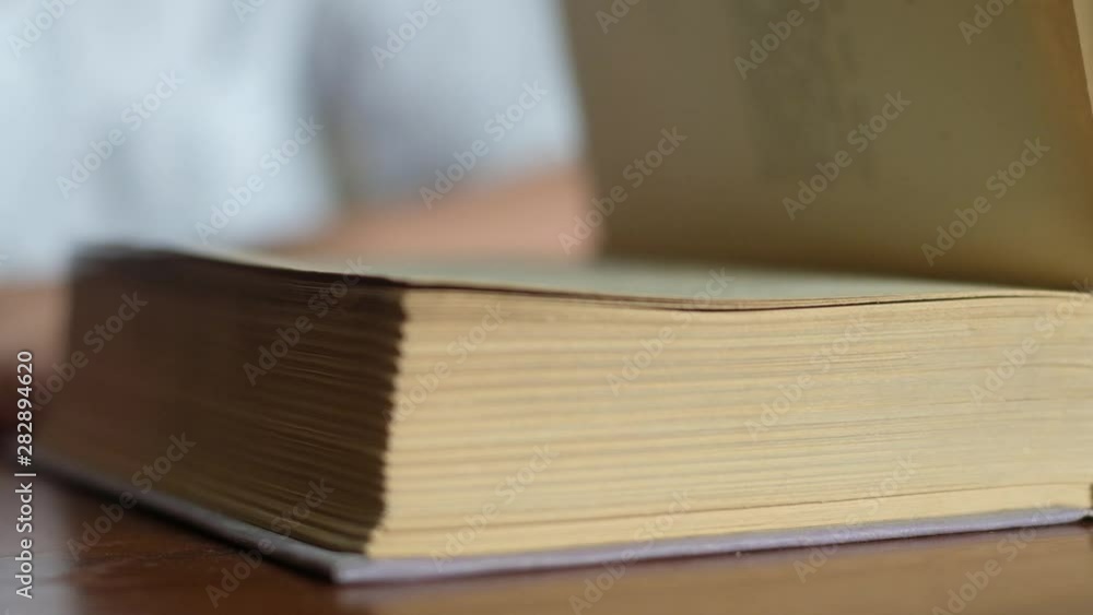 the student quickly turns the pages of a large book lying on a wooden table and closes the cover of literature. Close-up, angle view the student quickly turns the pages of a large book lying on a wooden table and closes the cover of literature. Close-up, angle view