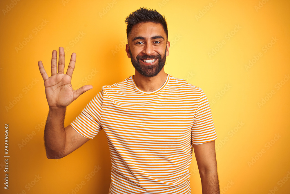 Young indian man wearing t-shirt standing over isolated yellow background showing and pointing up with fingers number five while smiling confident and happy.