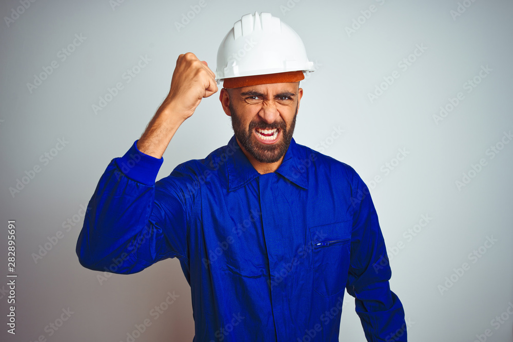 Handsome indian worker man wearing uniform and helmet over isolated white background angry and mad raising fist frustrated and furious while shouting with anger. Rage and aggressive concept.