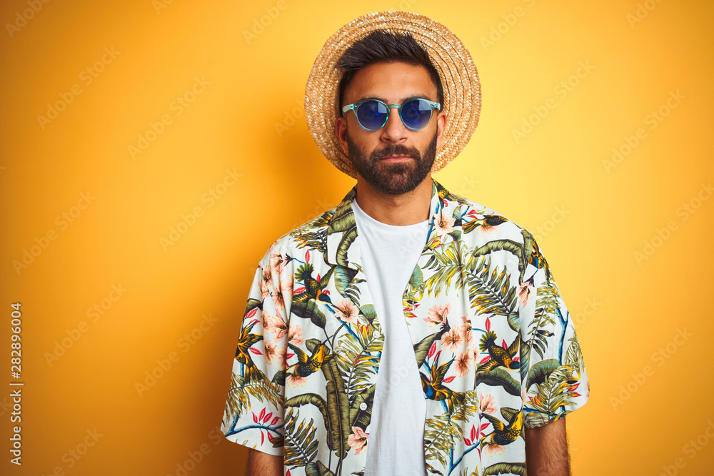Indian man on vacation wearing floral shirt hat sunglasses over isolated yellow background with serious expression on face. Simple and natural looking at the camera.