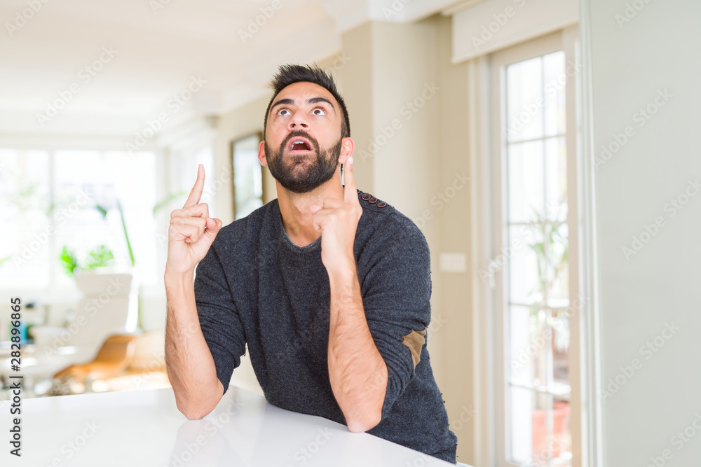 Handsome hispanic man wearing casual sweater at home amazed and surprised looking up and pointing with fingers and raised arms. Handsome hispanic man wearing casual sweater at home amazed and surprised looking up and pointing with fingers and raised arms.