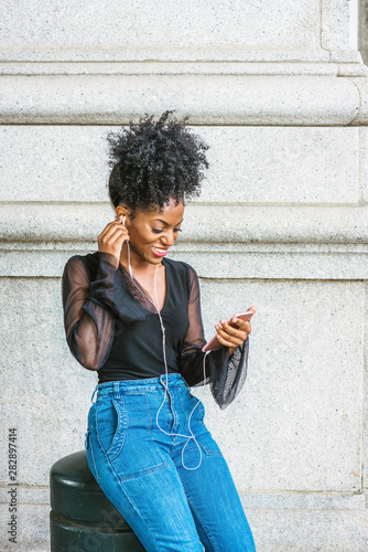 Young African American Woman with afro hairstyle, wearing mesh sheer long sleeve shirt blouse, blue jeans, sitting on street in New York City, listening music with earphone and cell phone, texting..