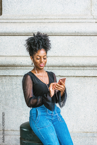 Young African American Woman with afro hairstyle, wearing mesh sheer long sleeve shirt blouse, blue jeans, sitting on street in New York City, listening music with earphone and cell phone, texting..