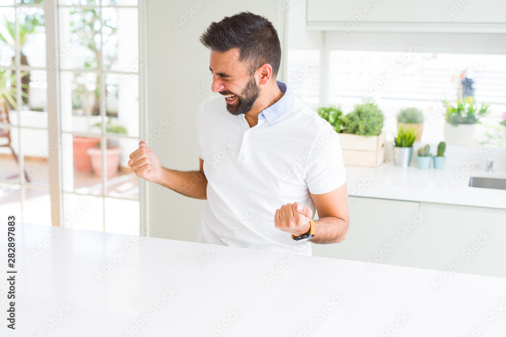 Handsome hispanic man casual white t-shirt at home very happy and excited doing winner gesture with arms raised, smiling and screaming for success. Celebration concept. Handsome hispanic man casual white t-shirt at home very happy and excited doing winner gesture with arms raised, smiling and screaming for success. Celebration concept.