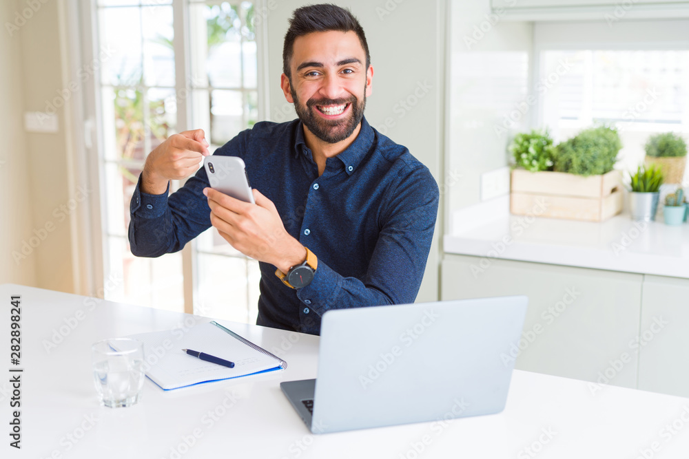 Handsome hispanic business man using smartphone and laptop at the office very happy pointing with hand and finger