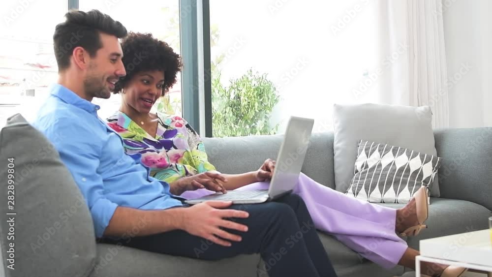 Content relaxed caucasian man and African american woman enjoying time together and browsing laptop on comfortable couch against window Content relaxed caucasian man and African american woman enjoying time together and browsing laptop on comfortable couch against window