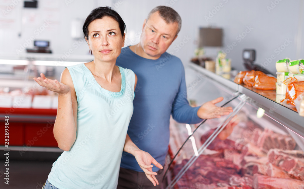 Sad adult couple customers choosing meat in shop
