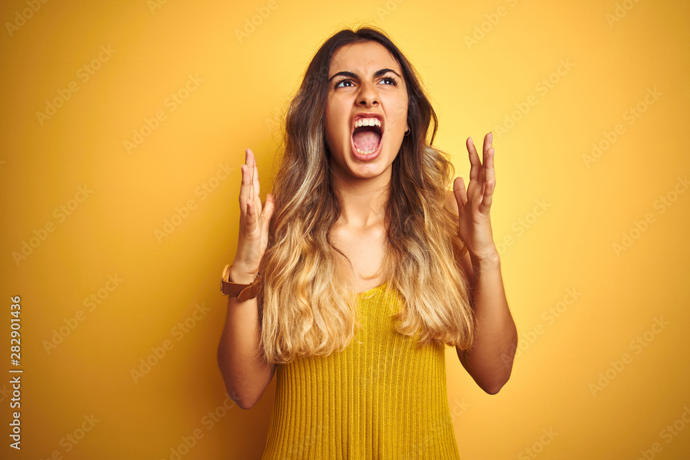 Young beautiful woman wearing t-shirt over yellow isolated background crazy and mad shouting and yelling with aggressive expression and arms raised. Frustration concept. Young beautiful woman wearing t-shirt over yellow isolated background crazy and mad shouting and yelling with aggressive expression and arms raised. Frustration concept.