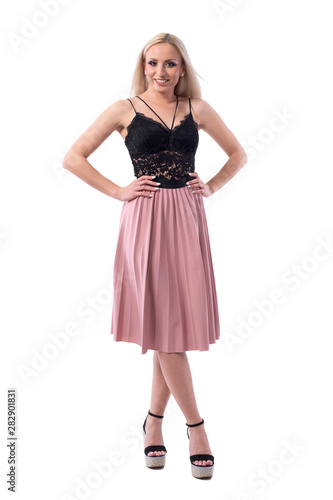 Cute pretty blonde woman in black lace top and salmon color skirt posing with arms on hips. Full body isolated on white background. 