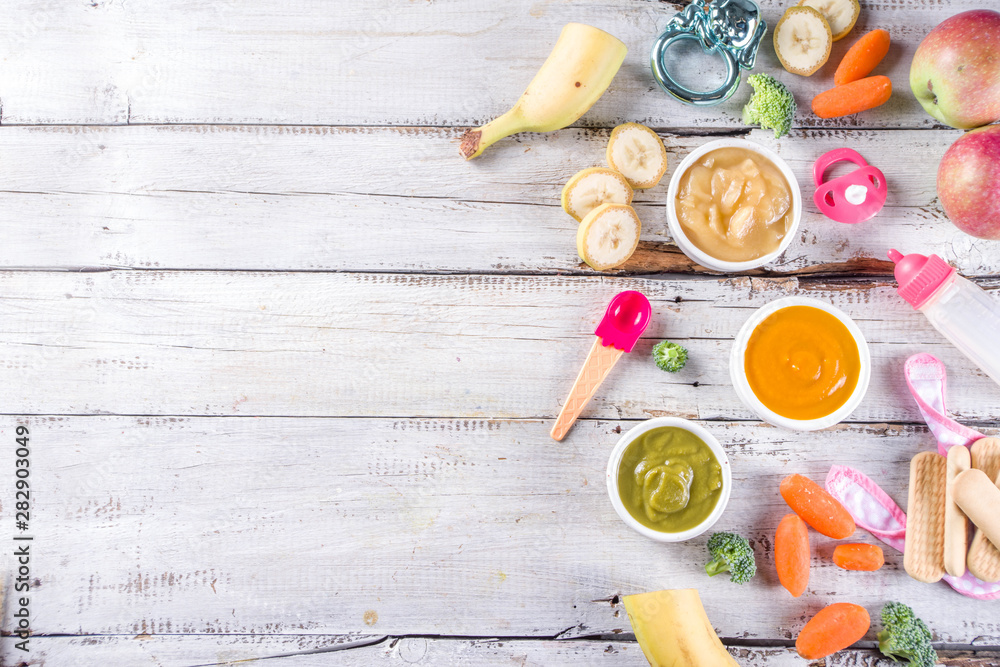 Infant baby food. Bowls with vegetable fruit puree, green, orange, yellow colors - broccoli, carrots, banana, apple. With baby accessories and toys wooden white background Flat lay top view Infant baby food. Bowls with vegetable fruit puree, green, orange, yellow colors - broccoli, carrots, banana, apple. With baby accessories and toys wooden white background Flat lay top view