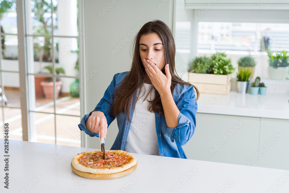 Beautiful young woman cutting a tasty pizza slice using a cutter cover mouth with hand shocked with shame for mistake, expression of fear, scared in silence, secret concept Beautiful young woman cutting a tasty pizza slice using a cutter cover mouth with hand shocked with shame for mistake, expression of fear, scared in silence, secret concept