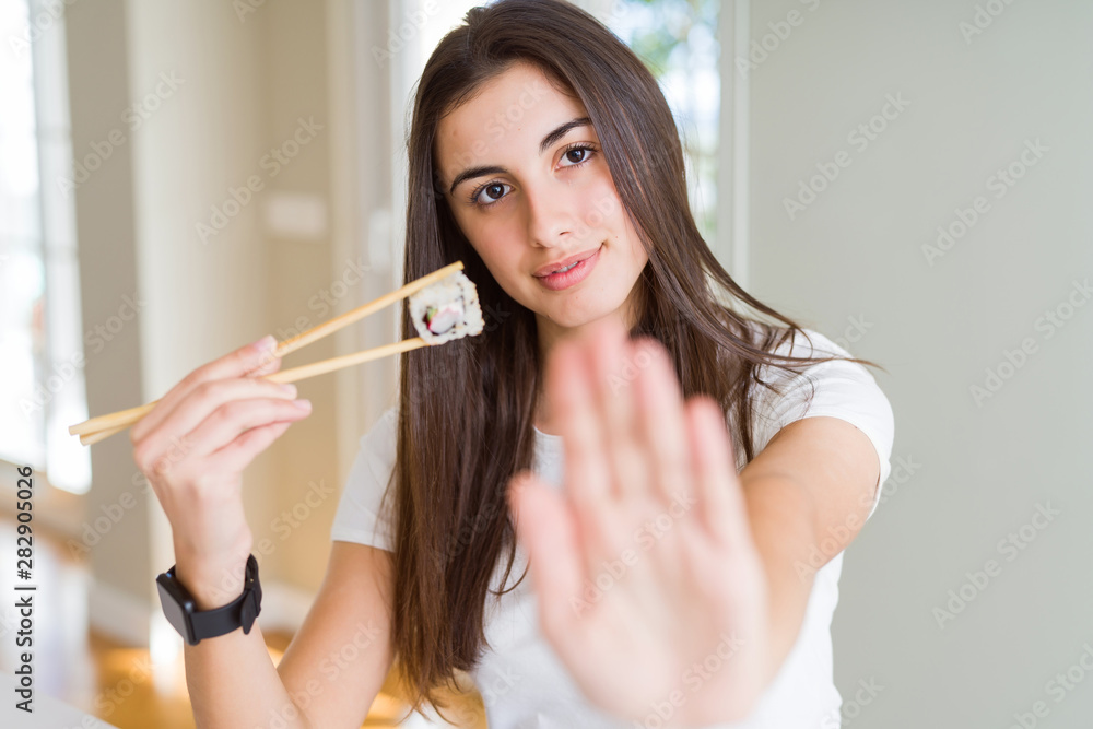 Beautiful young woman eating asian sushi using chopsticks with open hand doing stop sign with serious and confident expression, defense gesture Beautiful young woman eating asian sushi using chopsticks with open hand doing stop sign with serious and confident expression, defense gesture