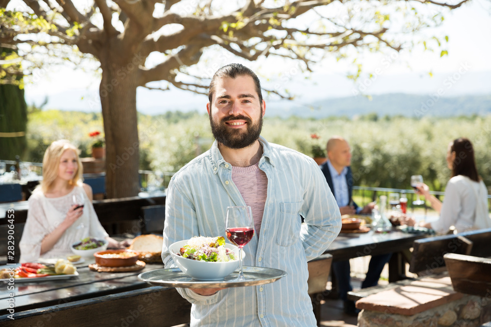 Adult beard waiter who is standing with order at country outdoor restaurant