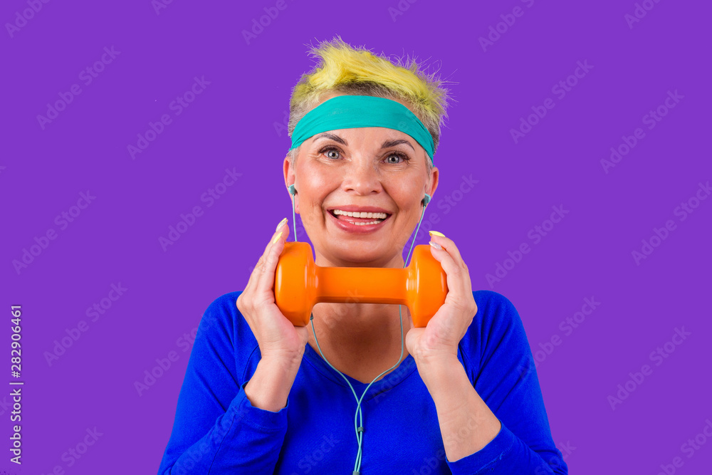 cheerful elderly female with stylish yellow dyed hair and a blue bandage on forehead training with orange dumbbelland listening to music on headphones in gym violet studio background cheerful elderly female with stylish yellow dyed hair and a blue bandage on forehead training with orange dumbbelland listening to music on headphones in gym violet studio background
