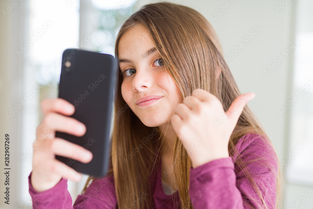 Beautiful young girl kid sending a message using smartphone pointing and showing with thumb up to the side with happy face smiling Beautiful young girl kid sending a message using smartphone pointing and showing with thumb up to the side with happy face smiling