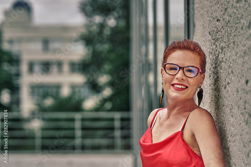 Portrait of a red-haired laughing middle-aged woman in a red blouse standing against the wall. Cloudy summer day. Close-up.