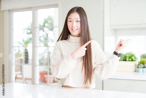 Beautiful Asian woman wearing casual sweater smiling and looking at the camera pointing with two hands and fingers to the side.