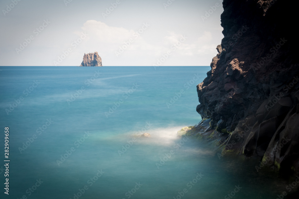 A light house on a small island, seen from a cliff with black rock.