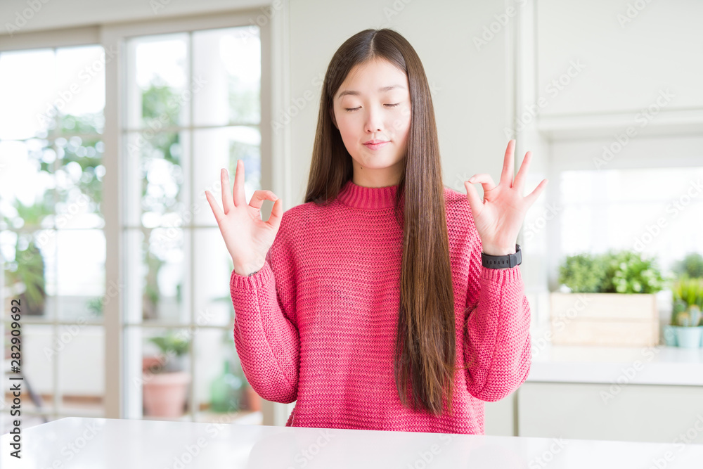 Beautiful Asian woman wearing pink sweater on white table relax and smiling with eyes closed doing meditation gesture with fingers. Yoga concept. Beautiful Asian woman wearing pink sweater on white table relax and smiling with eyes closed doing meditation gesture with fingers. Yoga concept.