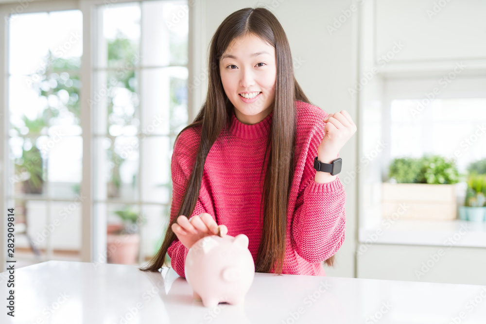 Beautiful Asian woman putting a coin inside piggy bag screaming proud and celebrating victory and success very excited, cheering emotion Beautiful Asian woman putting a coin inside piggy bag screaming proud and celebrating victory and success very excited, cheering emotion