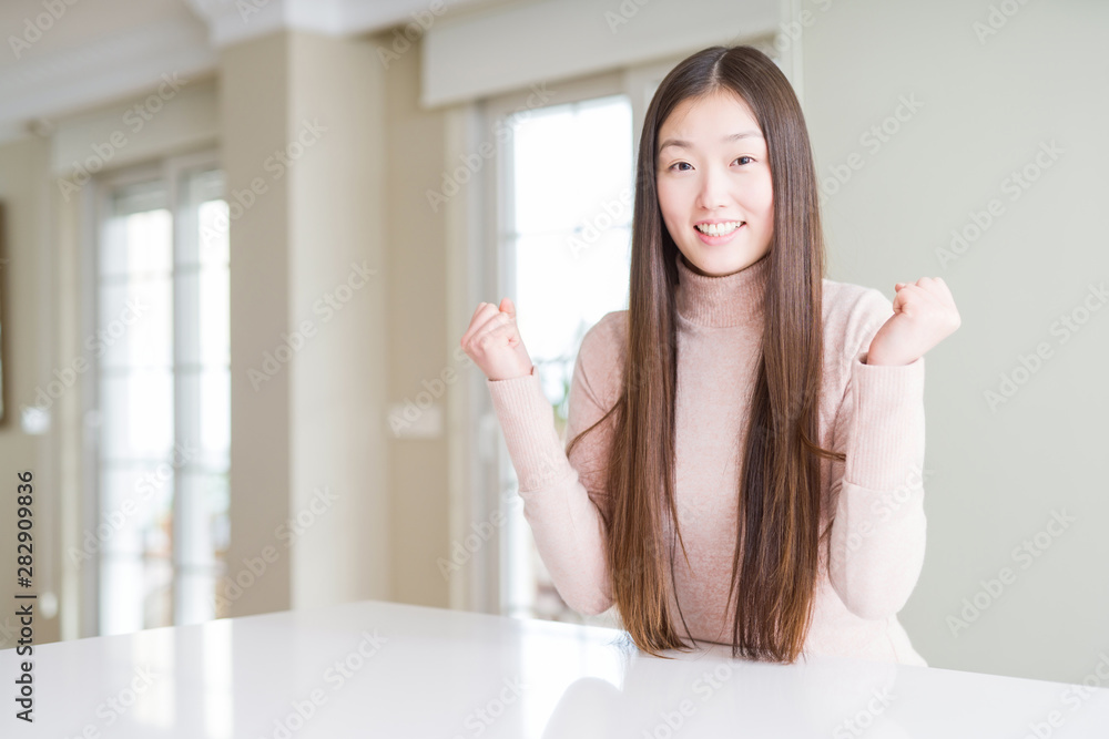 Beautiful Asian woman wearing casual sweater on white table celebrating surprised and amazed for success with arms raised and open eyes. Winner concept. Beautiful Asian woman wearing casual sweater on white table celebrating surprised and amazed for success with arms raised and open eyes. Winner concept.