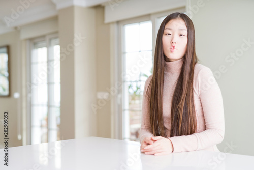 Beautiful Asian woman wearing casual sweater on white table making fish face with lips, crazy and comical gesture. Funny expression.