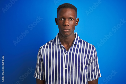 African american man wearing striped casual shirt standing over isolated blue background with serious expression on face. Simple and natural looking at the camera.