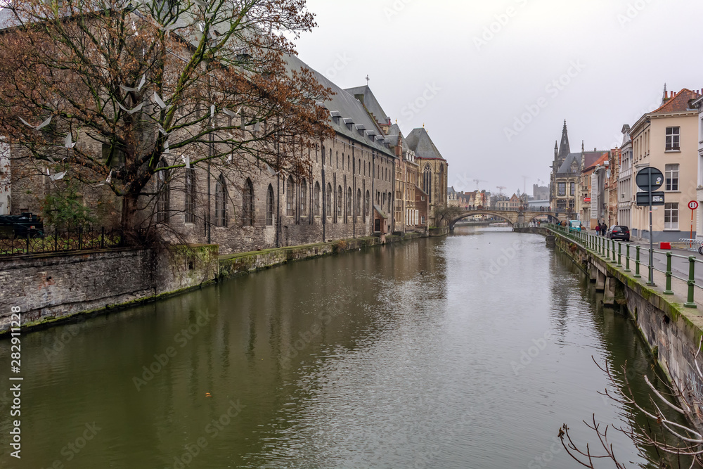 Old street of morning Ghent, Belgium, with traditional medieval houses, canal, bridge,  tower in the background and white paper birds on the tree in the foreground.