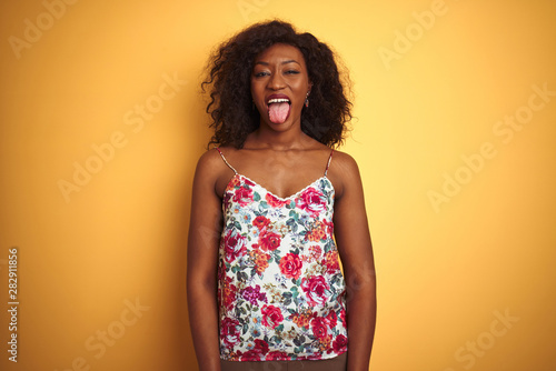 African american woman wearing floral summer t-shirt over isolated yellow background sticking tongue out happy with funny expression. Emotion concept.