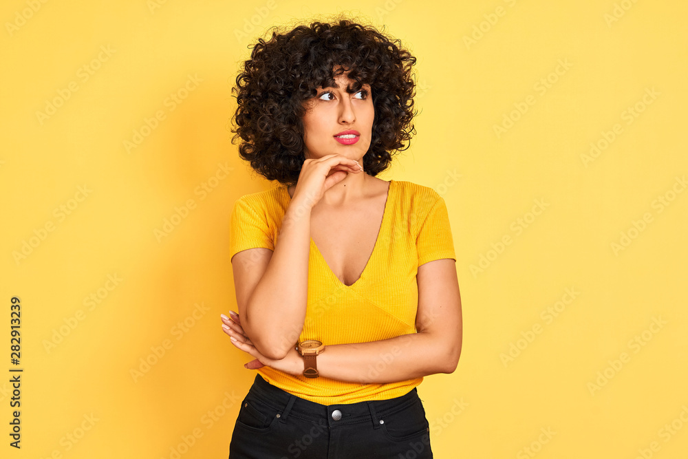Young arab woman with curly hair wearing t-shirt standing over isolated yellow background with hand on chin thinking about question, pensive expression. Smiling with thoughtful face. Doubt concept.