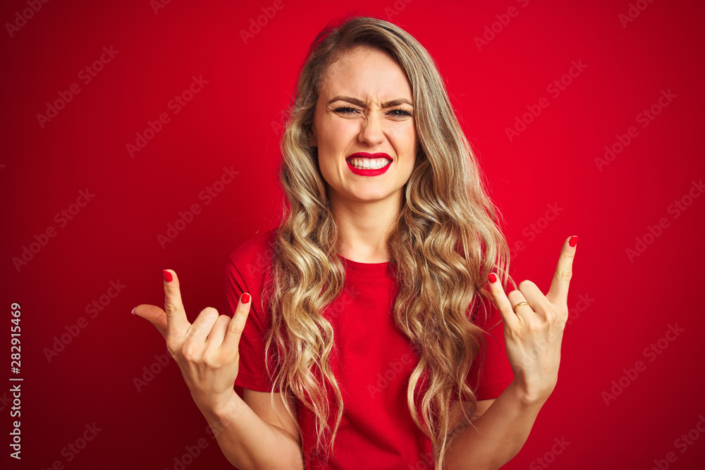 Young beautiful woman wearing basic t-shirt standing over red isolated background shouting with crazy expression doing rock symbol with hands up. Music star. Heavy concept. Young beautiful woman wearing basic t-shirt standing over red isolated background shouting with crazy expression doing rock symbol with hands up. Music star. Heavy concept.