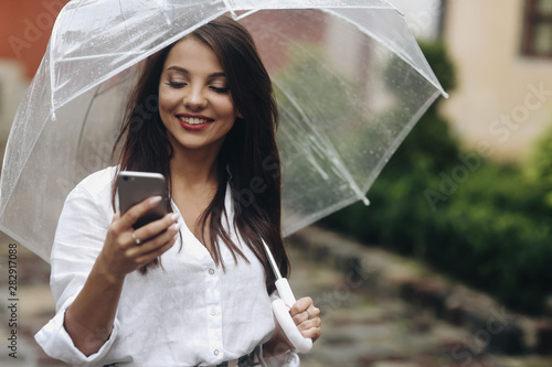 Portrait of smiling beautiful girl with umbrella calls a friend standing on the old street. It's summer rain.