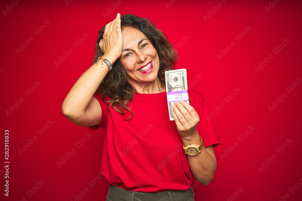 Middle age senior woman holding bunch of dollars over red isolated background stressed with hand on head, shocked with shame and surprise face, angry and frustrated. Fear and upset for mistake. Middle age senior woman holding bunch of dollars over red isolated background stressed with hand on head, shocked with shame and surprise face, angry and frustrated. Fear and upset for mistake.