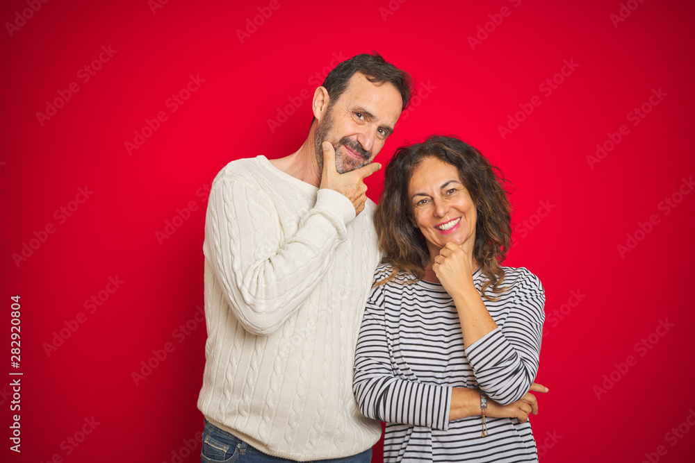 Beautiful middle age couple wearing winter sweater over isolated red background looking confident at the camera smiling with crossed arms and hand raised on chin. Thinking positive. Beautiful middle age couple wearing winter sweater over isolated red background looking confident at the camera smiling with crossed arms and hand raised on chin. Thinking positive.