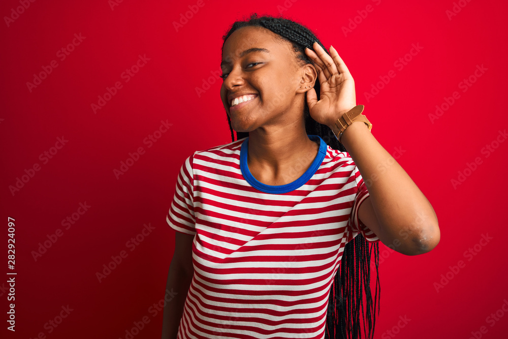 Young african american woman wearing striped t-shirt standing over isolated red background smiling with hand over ear listening an hearing to rumor or gossip. Deafness concept. Young african american woman wearing striped t-shirt standing over isolated red background smiling with hand over ear listening an hearing to rumor or gossip. Deafness concept.