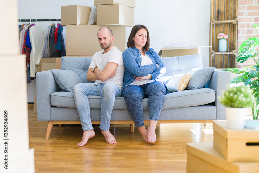 Young couple sitting on the sofa arround cardboard boxes moving to a new house skeptic and nervous, disapproving expression on face with crossed arms. Negative person. Young couple sitting on the sofa arround cardboard boxes moving to a new house skeptic and nervous, disapproving expression on face with crossed arms. Negative person.