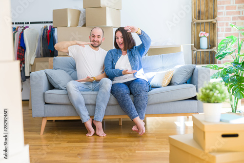 Young couple sitting on the sofa arround cardboard boxes moving to a new house gesturing with hands showing big and large size sign, measure symbol. Smiling looking at the camera. Measuring concept. © Krakenimages.com