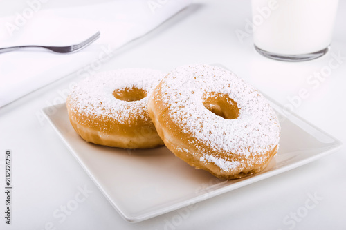 Donuts covered with icing sugar on a plate on a white background accompanied by a glass of milk and a fork on a napkin. Delicious breakfast or lunch. American food. Bakery and pastry products.