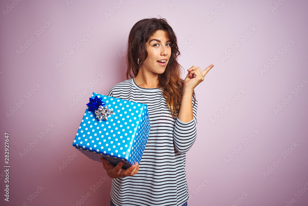 Young beautiful woman holding birthday gift over pink isolated background very happy pointing with hand and finger to the side Young beautiful woman holding birthday gift over pink isolated background very happy pointing with hand and finger to the side