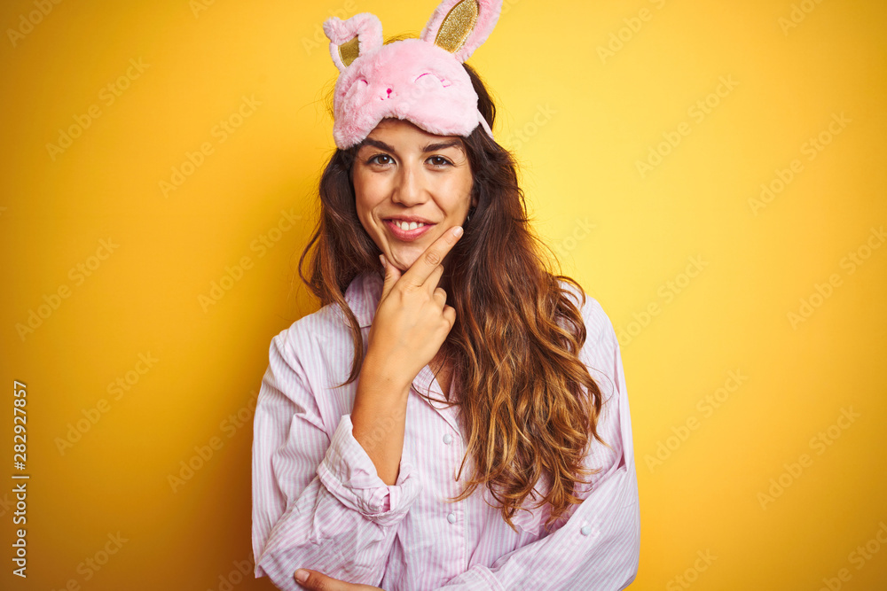 Young woman wearing pajama and sleep mask standing over yellow isolated background looking confident at the camera smiling with crossed arms and hand raised on chin. Thinking positive. Young woman wearing pajama and sleep mask standing over yellow isolated background looking confident at the camera smiling with crossed arms and hand raised on chin. Thinking positive.