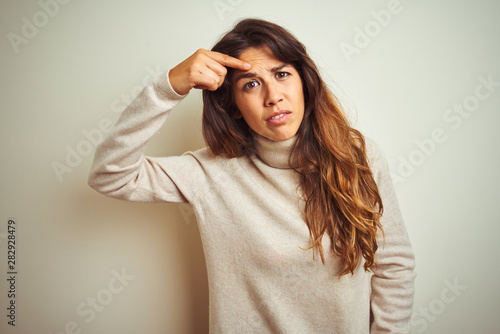 Young beautiful woman wearing winter sweater standing over white isolated background pointing unhappy to pimple on forehead, ugly infection of blackhead. Acne and skin problem