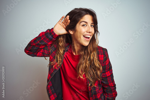 Young beautiful woman wearing red t-shirt and jacket standing over white isolated background smiling with hand over ear listening an hearing to rumor or gossip. Deafness concept.