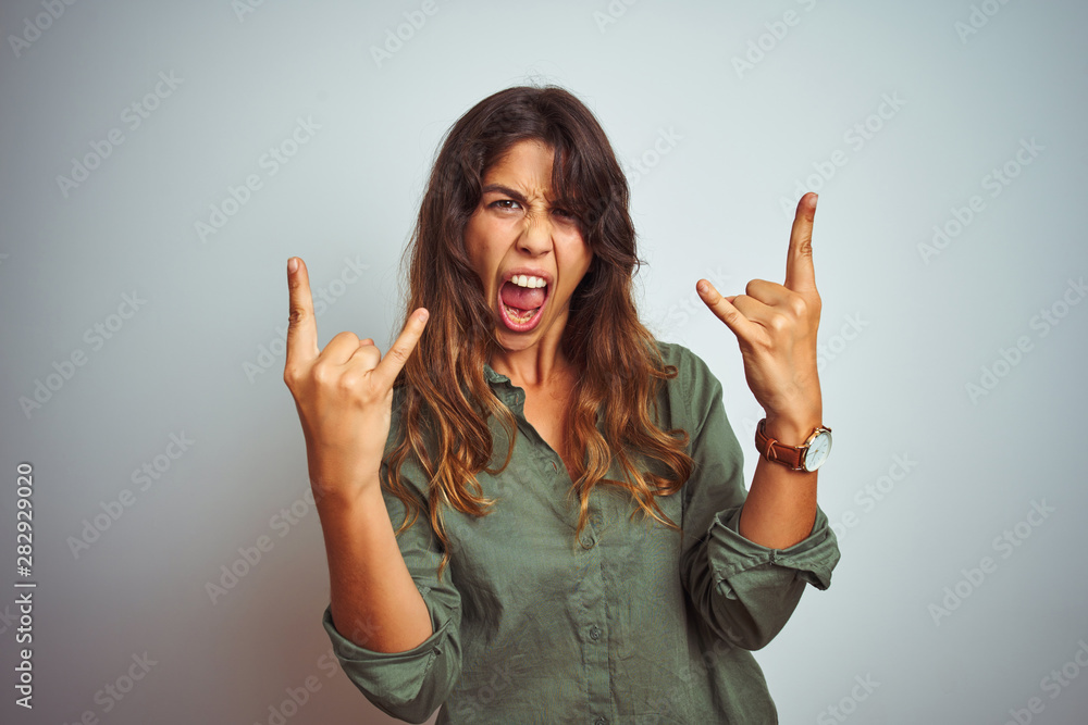Young beautiful woman wearing green shirt standing over grey isolated background shouting with crazy expression doing rock symbol with hands up. Music star. Heavy music concept. Young beautiful woman wearing green shirt standing over grey isolated background shouting with crazy expression doing rock symbol with hands up. Music star. Heavy music concept.