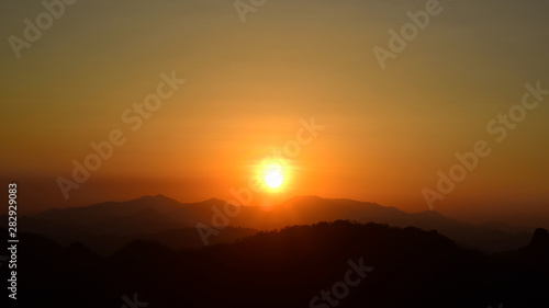 sun setting behind a dense forest area followed by mountains. © tharathip