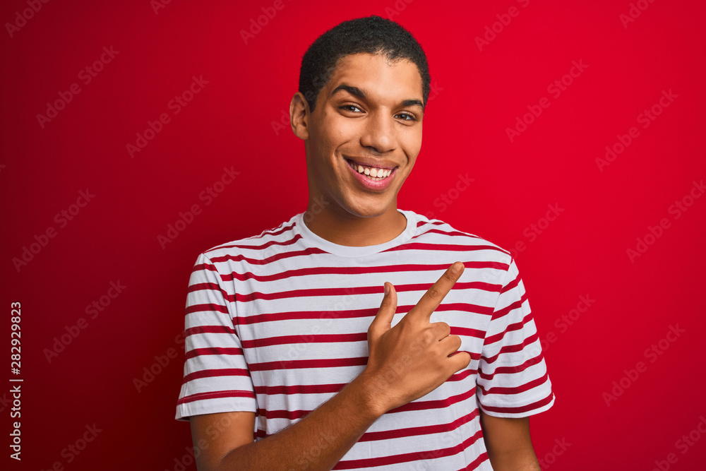 Young handsome arab man wearing striped t-shirt over isolated red background cheerful with a smile on face pointing with hand and finger up to the side with happy and natural expression Young handsome arab man wearing striped t-shirt over isolated red background cheerful with a smile on face pointing with hand and finger up to the side with happy and natural expression