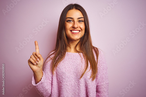 Young beautiful woman wearing casual sweater standing over isolated pink background showing and pointing up with finger number one while smiling confident and happy.