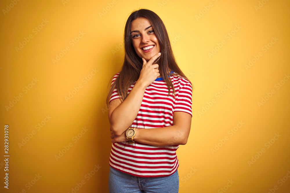 Young beautiful woman wearing striped t-shirt standing over isolated yellow background looking confident at the camera smiling with crossed arms and hand raised on chin. Thinking positive. Young beautiful woman wearing striped t-shirt standing over isolated yellow background looking confident at the camera smiling with crossed arms and hand raised on chin. Thinking positive.