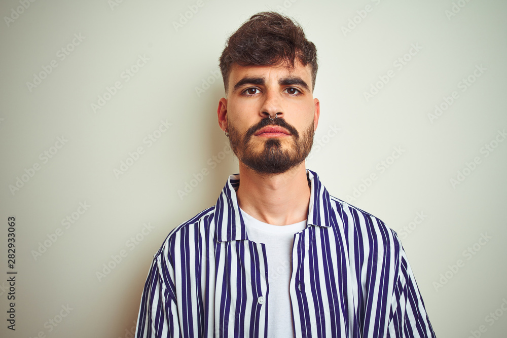 Young man with tattoo wearing striped shirt standing over isolated white background with serious expression on face. Simple and natural looking at the camera. Young man with tattoo wearing striped shirt standing over isolated white background with serious expression on face. Simple and natural looking at the camera.