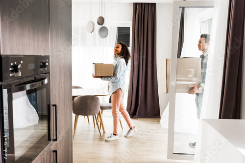 side view of african american husband and wife holding boxes while walking into house