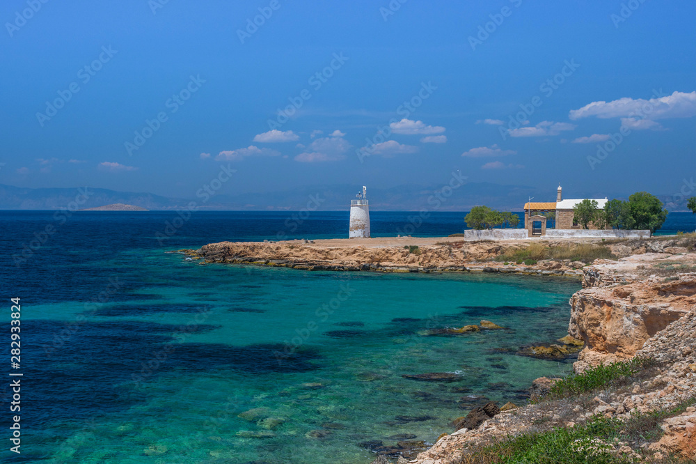 The wild coast of Aegina island with clear and blue waters of Mediterranean sea and the old small lighthouse in the background, in Saronic gulf, Greece.